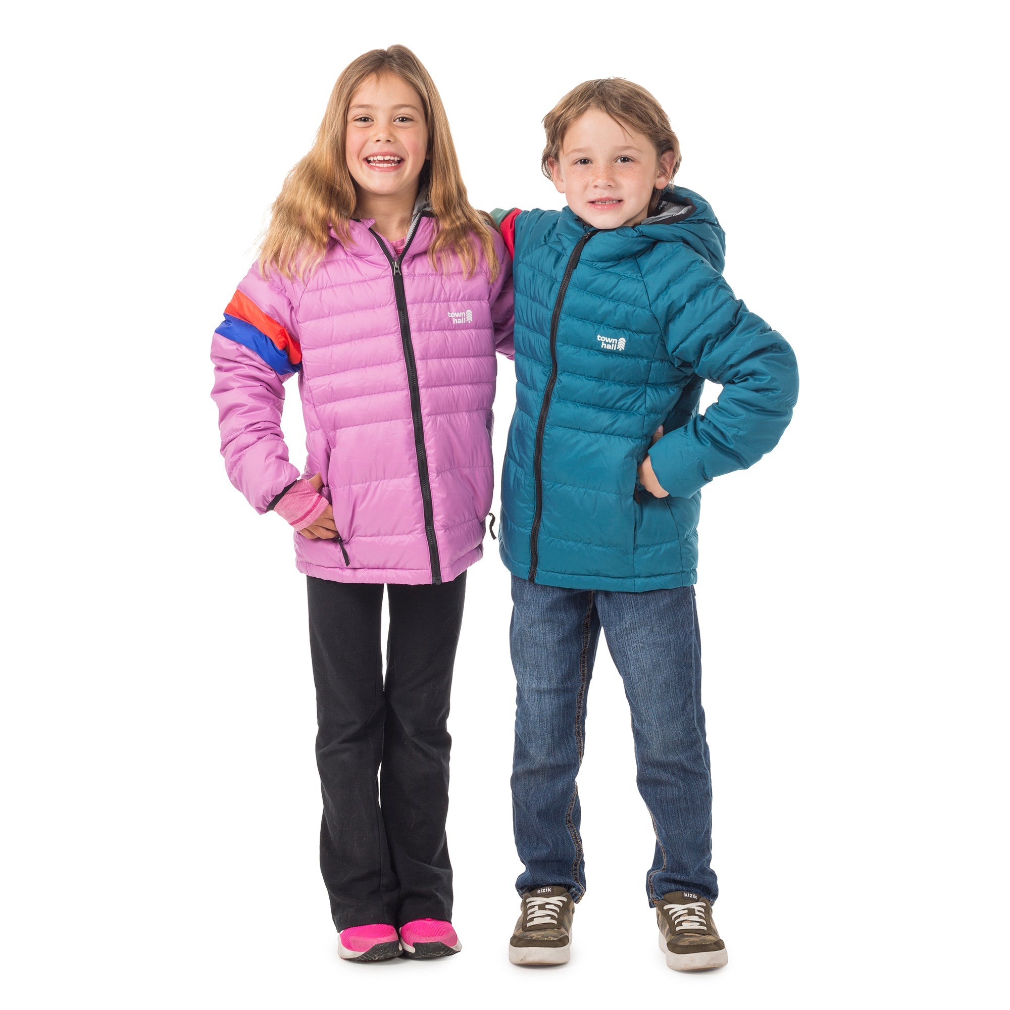 Two smiling children stand together. The girl wears the Down Town Kids Puffy jacket in pink with black pants and pink shoes, while the boy, dressed in a blue sustainable winter coat and jeans, wraps his arm around her as they grin.