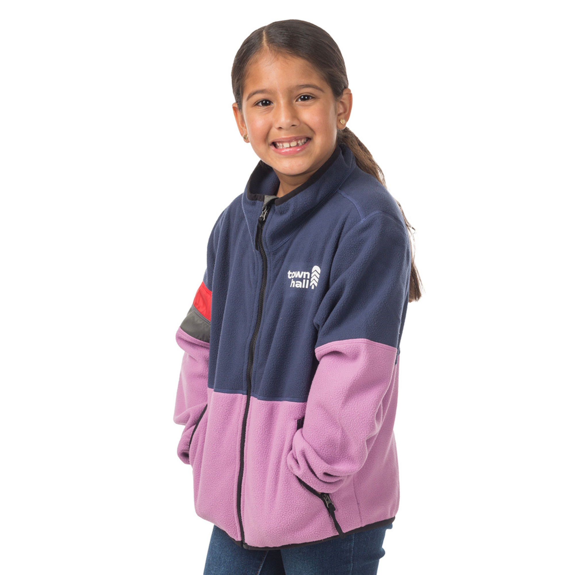 A young girl with long dark hair smiles, wearing a Midtown Fleece Jacket made from recycled polyester and featuring "twm hall" on the chest, standing with her hands in her pockets against a white background.