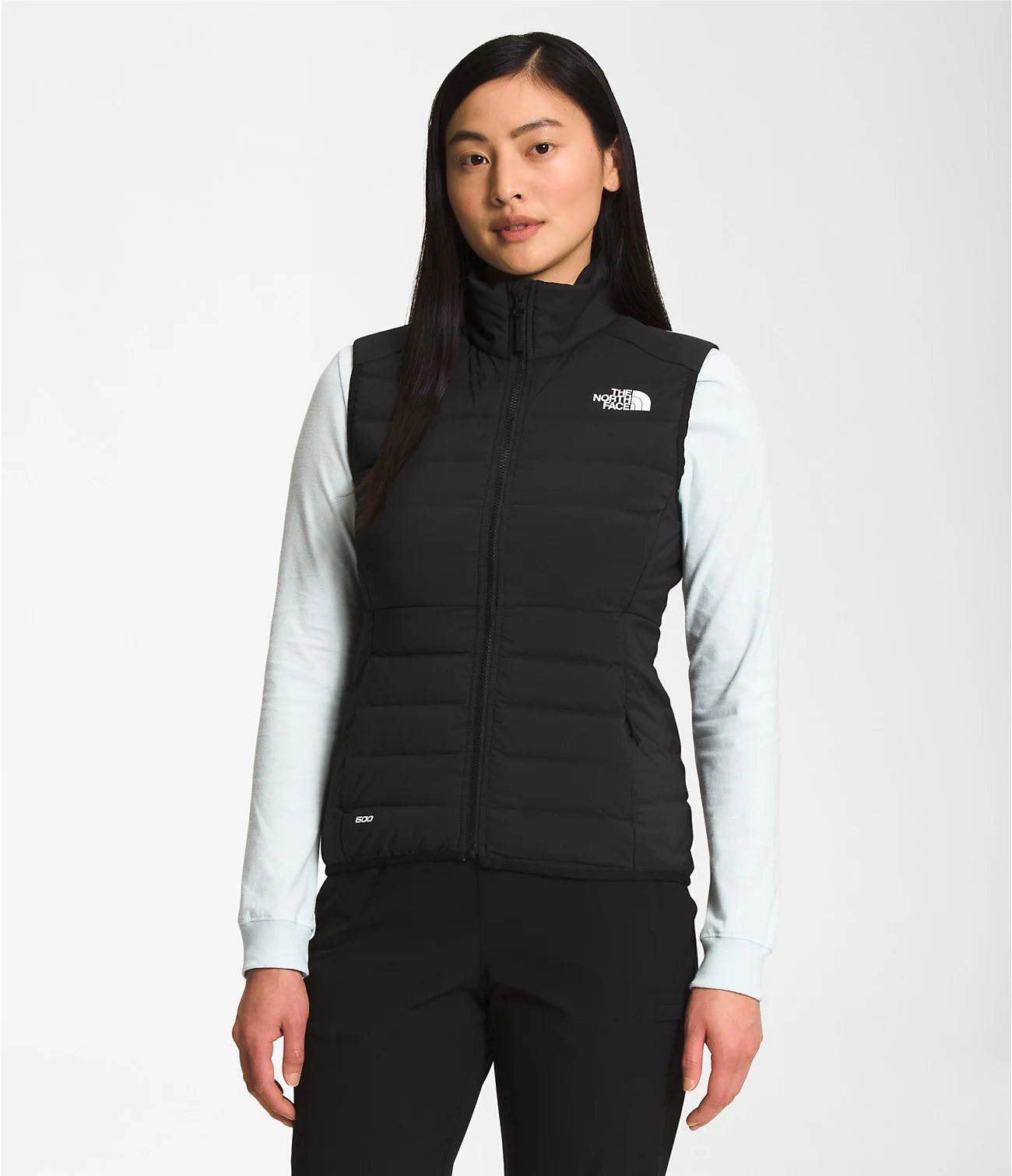 A woman wears The North Face Womens Belleview Stretch Down Vest—black, slim fit, sleeveless—over a light long-sleeve shirt, standing against a plain white background. This packable vest is ideal for layering on any adventure.