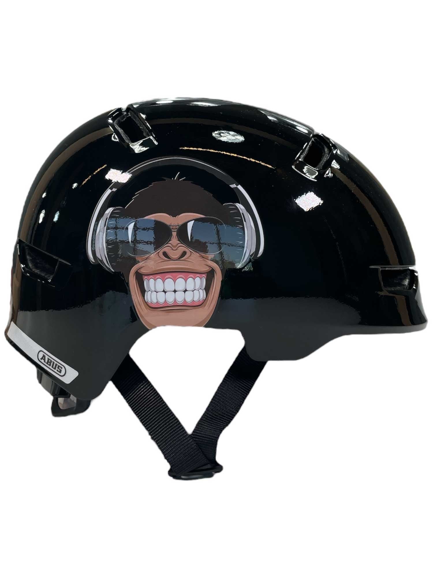 The Abus Kids' Scraper 3.0 Monkey Helmet is a shiny black helmet featuring a grinning chimpanzee with sunglasses, an adjustable black chin strap, top ventilation holes, and ponytail compatibility for comfort.