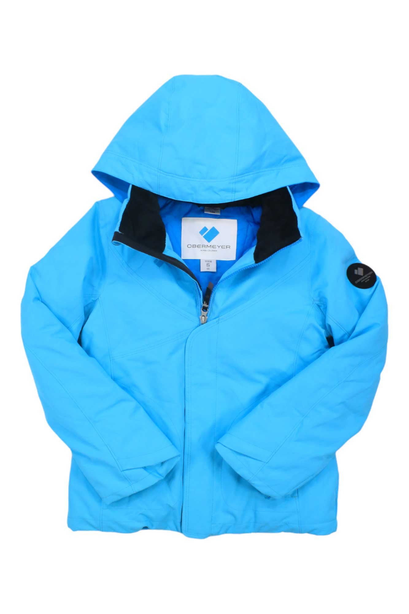A bright blue Obermeyer Girls Rylee Jacket with a front zipper, black inner collar, and a circular black patch on the left sleeve. This stylish ski jacket is laid flat, sleeves crossed—perfect for any kids’ outdoor adventure.