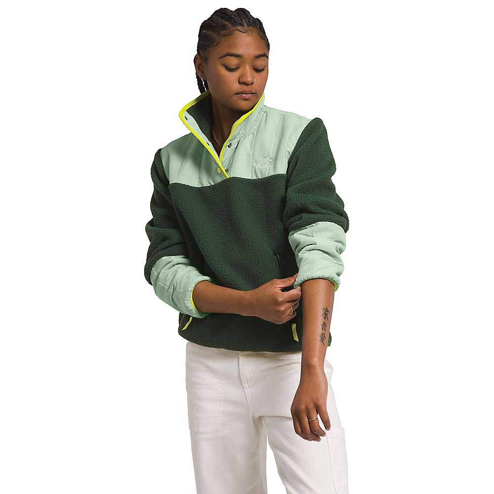 A person with braided hair wears a green The North Face Womens Cragmont Fleece 1/4 Snap Top and white pants, rolling up their sleeve to reveal a forearm tattoo.