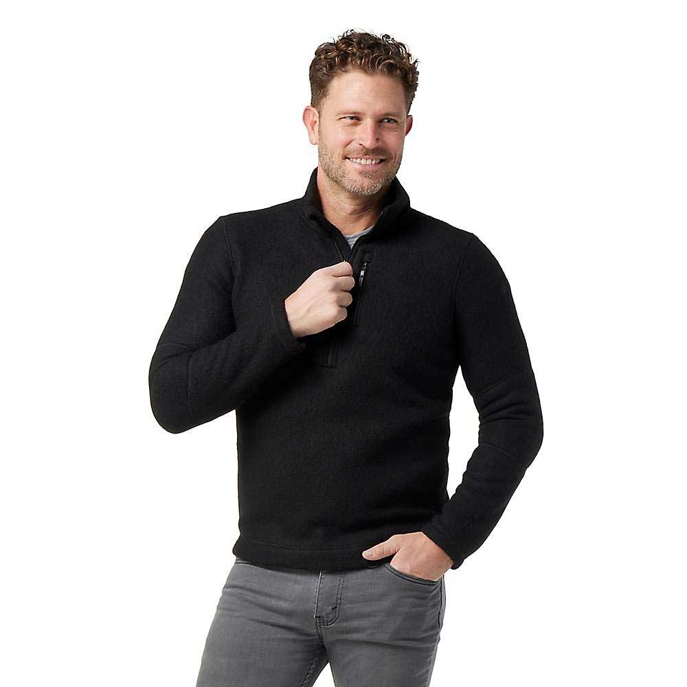 A man with curly hair and a trimmed beard smiles while wearing the Smartwool Men's Hudson Trail Fleece Half Zip Sweater in black and grey jeans, standing against a plain white background.