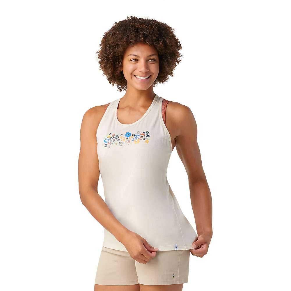 A woman with curly hair smiles, wearing the Smartwool Womens Floral Meadow Graphic Tank in light beige and tan shorts. She holds the hem of her moisture-wicking top with one hand against a plain white background.