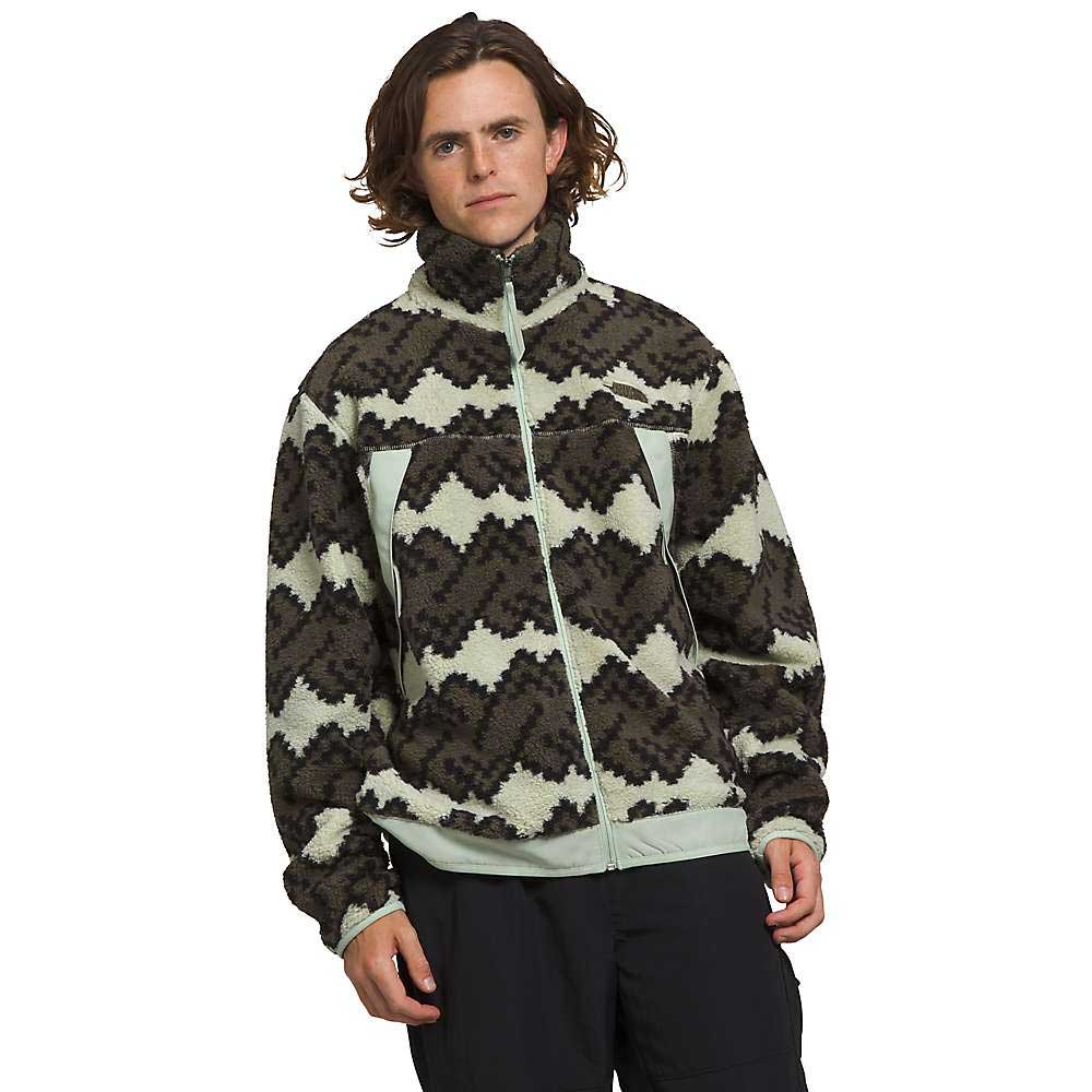 A person with shoulder-length brown hair faces forward in The North Face Mens Campshire Fleece Jacket, which displays black, gray, and cream zigzag shapes. They have a neutral expression and stand against a white background.