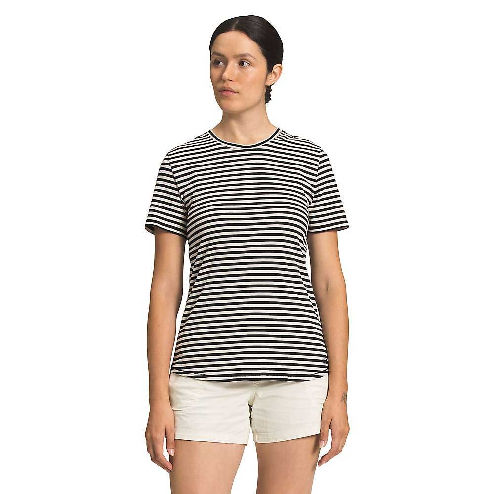 A woman stands against a plain white background, wearing The North Face Womens Best Tee Ever in black and white stripes—a women's slim fit t-shirt made from organic cotton—paired with light shorts. Her dark hair is pulled back as she looks slightly to the side.