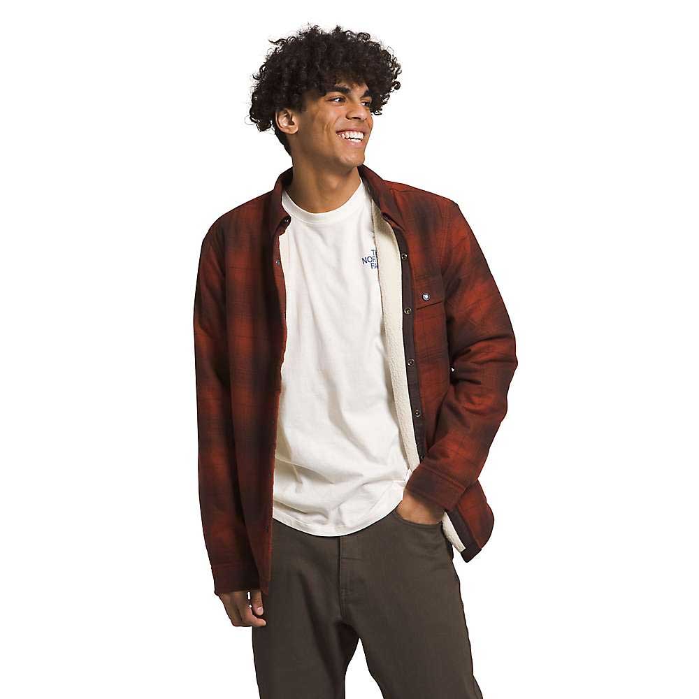 A young man with curly hair smiles while wearing The North Face Men's Campshire Shirt over a white t-shirt and dark pants, standing casually with one hand in his pocket against a plain white background.