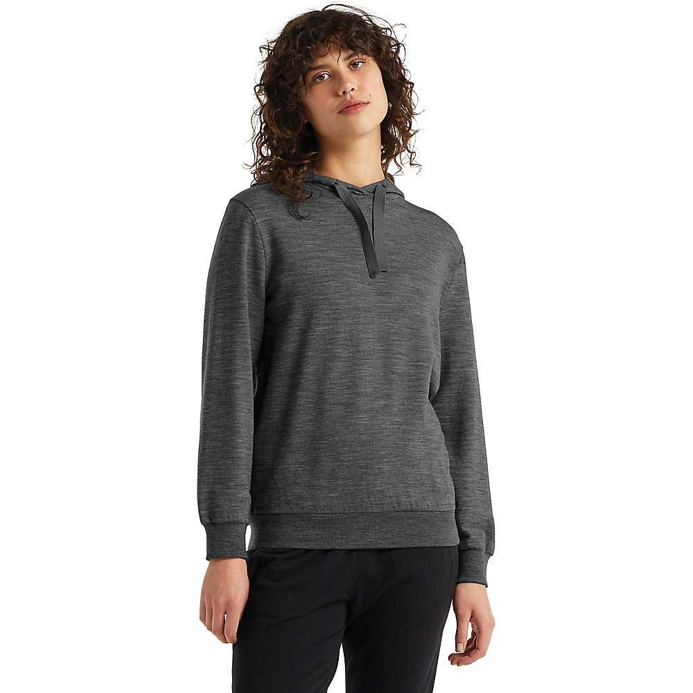 A woman with curly brown hair wears the Icebreaker Womens Crush LS Hoodie in dark gray and black pants, standing facing forward against a plain white background.