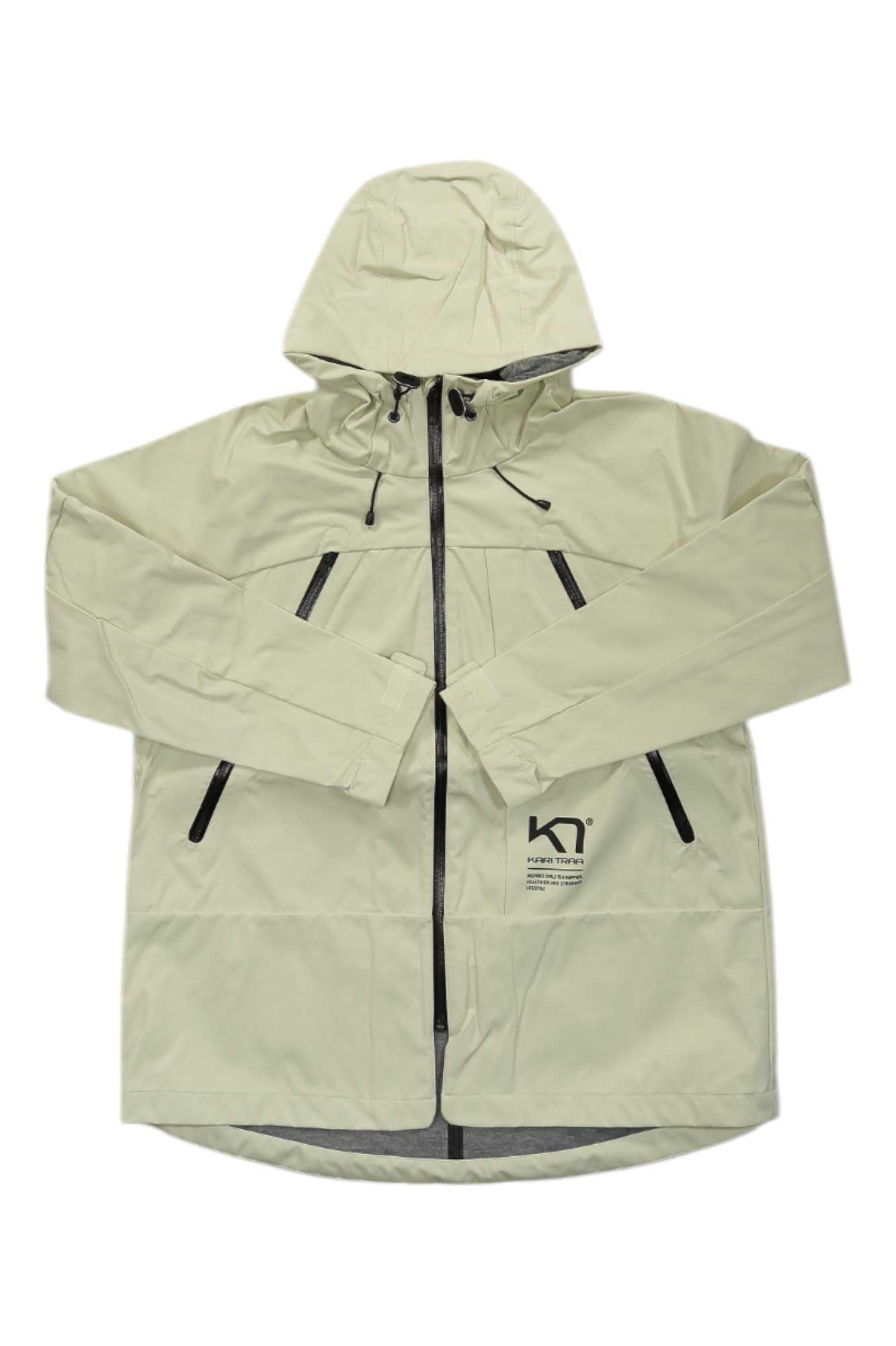 The Kari Traa Womens Bavallen Jacket is a light beige, wind-resistant jacket with black zipper closures on the front and chest pockets, shown flat on a white background. A small black logo and text label are on the lower left side.