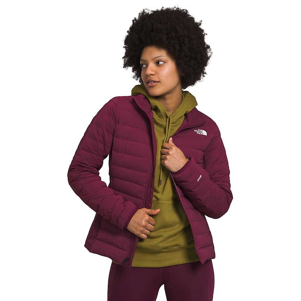 A person with curly hair wears The North Face Womens Belleview Stretch Down Jacket in maroon over an olive hoodie and matching pants, standing with one hand on the zipper and looking slightly to the side.