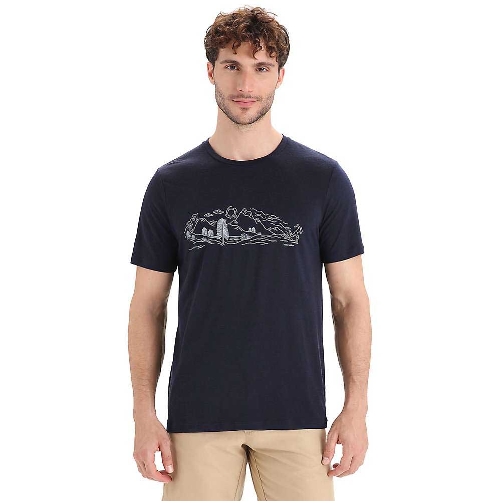 A man wears the Icebreaker Men's Tech Lite II SS Tee - Nature Sprint, featuring a navy blue short sleeve design with a white mountain, trees, and river graphic, paired with beige pants against a plain white background.