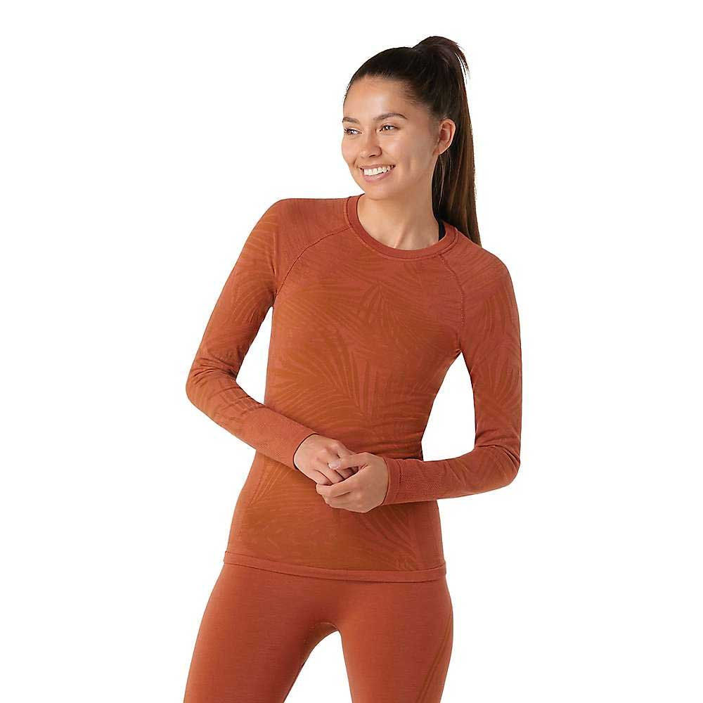 A woman with long brown hair in a ponytail smiles while wearing a rust-colored Smartwool Womens Intraknit Active Base Layer LS Top and leggings, standing against a plain white background.