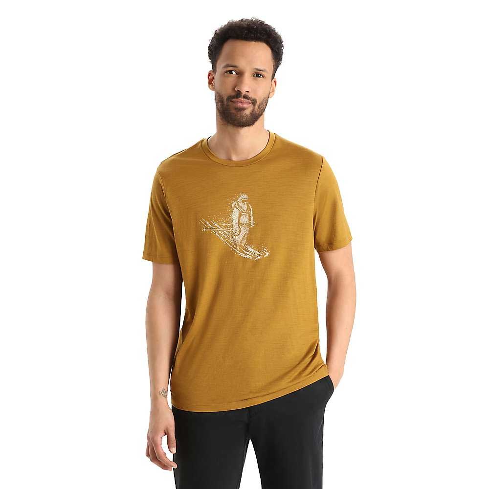 A man wearing the Icebreaker Mens Tech Lite II SS Tee - Skiing Yeti, featuring a mustard yellow color and a white skier graphic on the front, stands against a white background with one hand in his pocket.