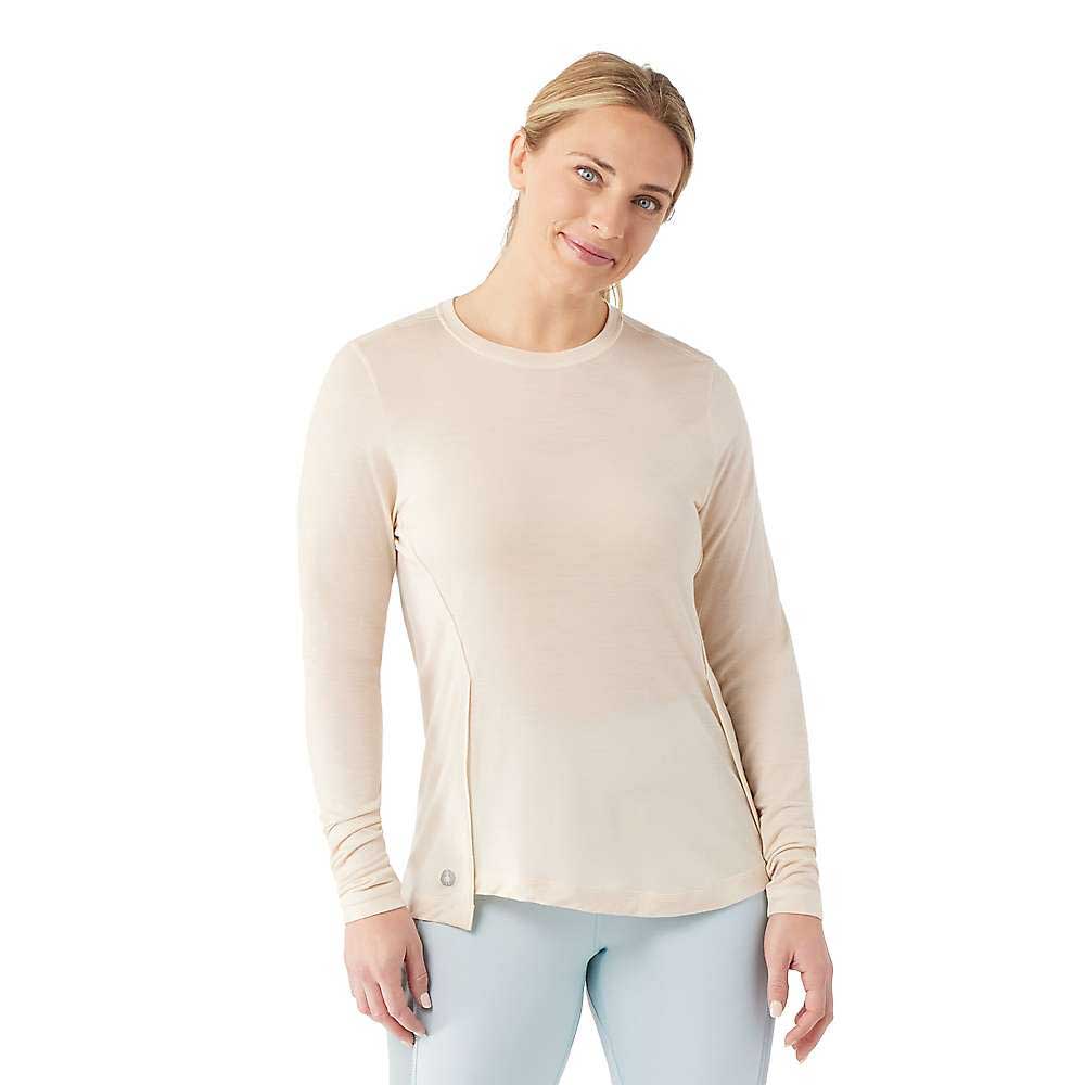 A woman with blonde hair tied back is wearing a Smartwool Womens Active LS Crew Top in light beige Merino wool and light blue pants, standing against a white background and smiling slightly.