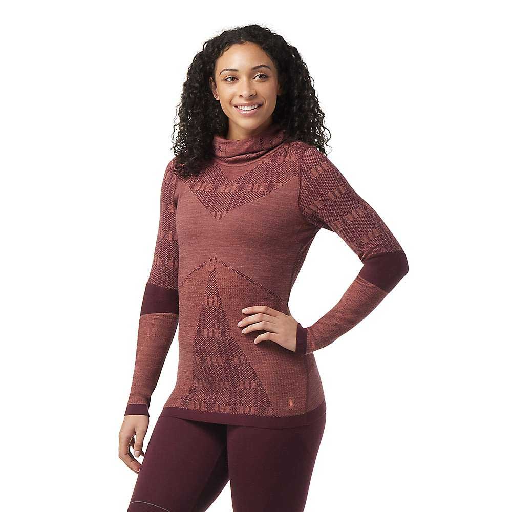 A woman with curly hair stands smiling, one hand on her hip, wearing the Smartwool Women's Intraknit Thermal Max Merino Base Layer Pattern Hoodie in maroon. The top has a high neck and moisture-wicking fabric for comfortable all-day wear.
