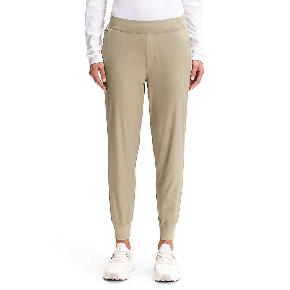 A person wearing The North Face Womens Aphrodite Jogger in light khaki, a white long-sleeve shirt, and white sneakers stands against a plain white background, shown from the shoulders down.