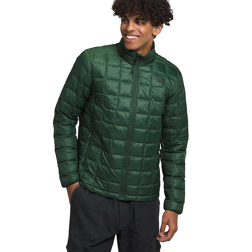 A young man with curly hair stands smiling to the side, wearing The North Face Men's ThermoBall Eco Jacket in dark green and dark pants. The jacket is made from recycled nylon. He poses against a plain white background.