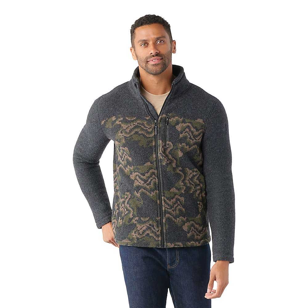 A man wears the Smartwool Men's Hudson Trail Fleece Full Zip Jacket—gray with a green and brown camo chest pattern, made from recycled wool—as he stands against a plain white background, looking forward with a slight smile.
