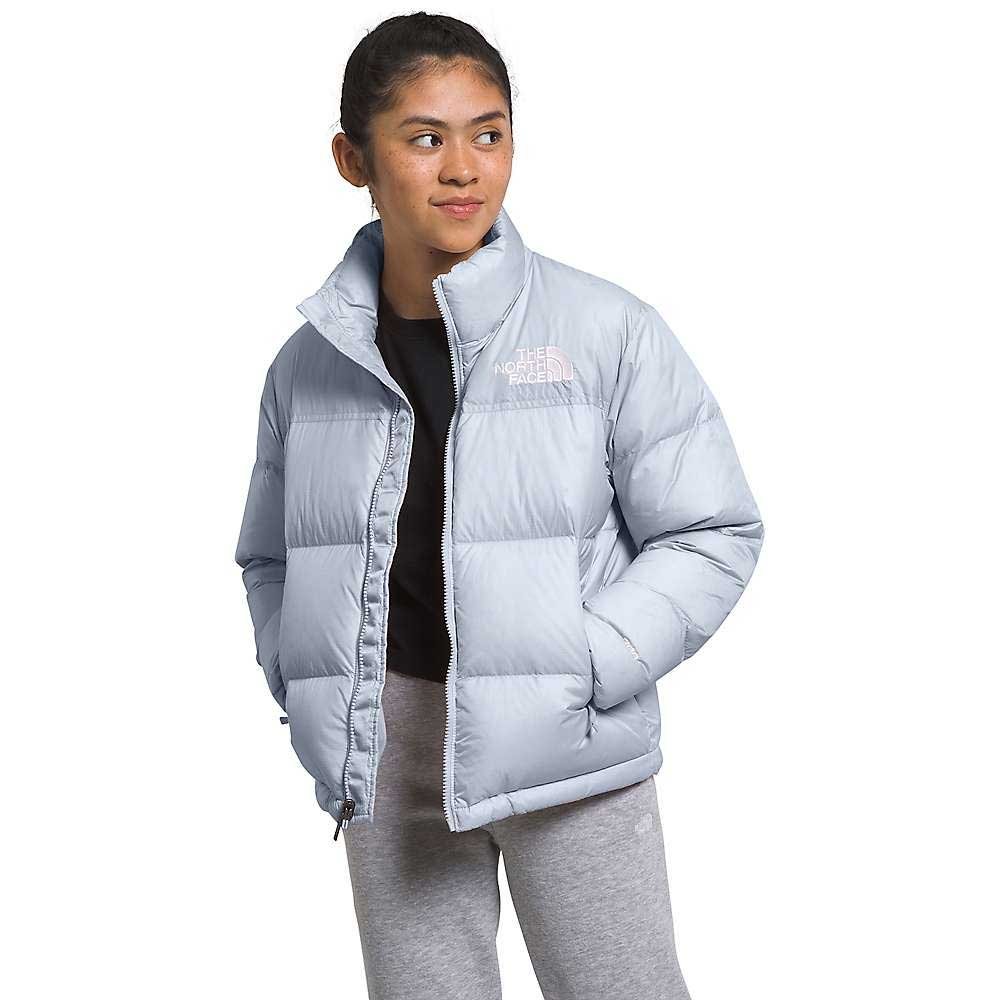 A young woman stands with her hands in her pockets, wearing The North Face Kids 1996 Retro Nuptse Jacket in light gray and gray sweatpants. This insulated jacket features the chest logo and is ideal for outdoor adventures.