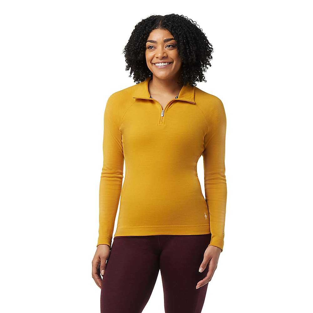 A person with curly hair smiles while wearing a mustard yellow Smartwool Women's Classic Thermal Merino Base Layer 1/4 Zip and dark maroon pants, standing against a plain white background.