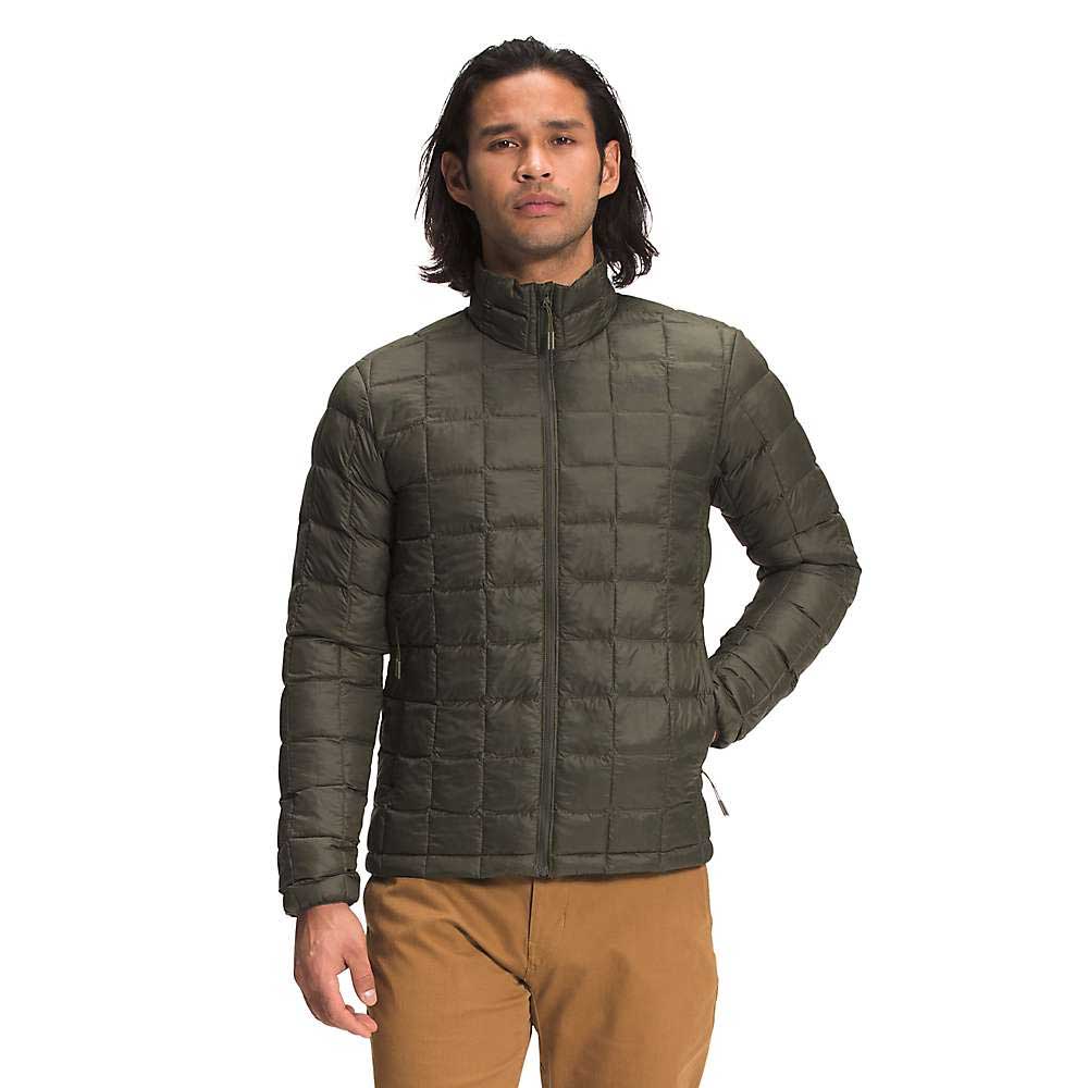 A person with long dark hair wears The North Face Men's ThermoBall Eco Jacket in olive green, made from recycled nylon, along with tan pants, standing and facing the camera against a plain white background.