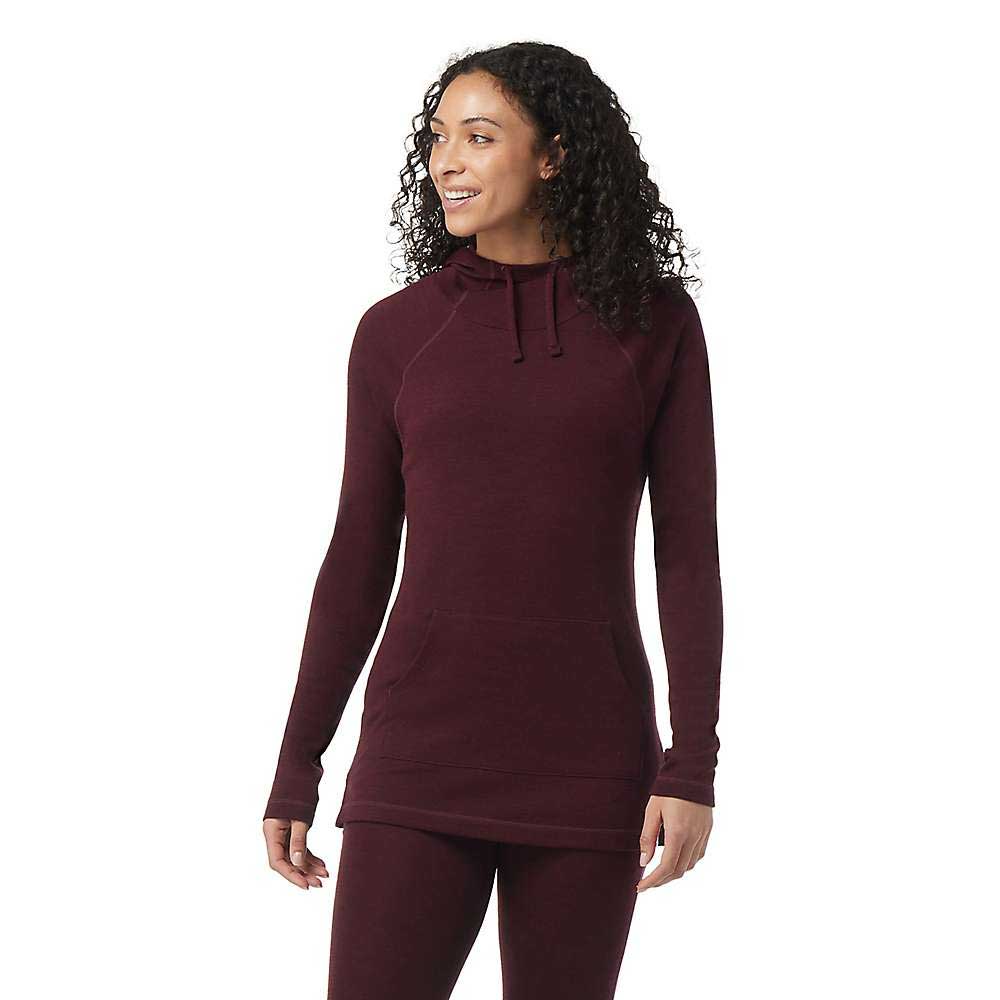 A woman with curly hair stands smiling, wearing the Smartwool Womens Thermal Merino Drape Neck Hoodie and matching pants, her hands relaxed by her sides as she faces slightly to the side.