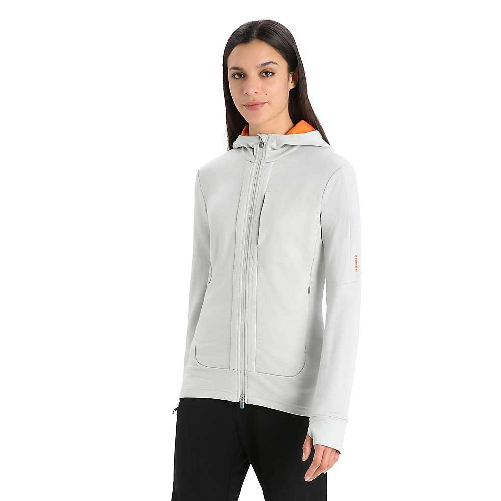 A woman stands slightly turned, wearing the Icebreaker Womens Quantum III LS Zip Hoodie in light gray with an orange lining and black pants, set against a plain white background.