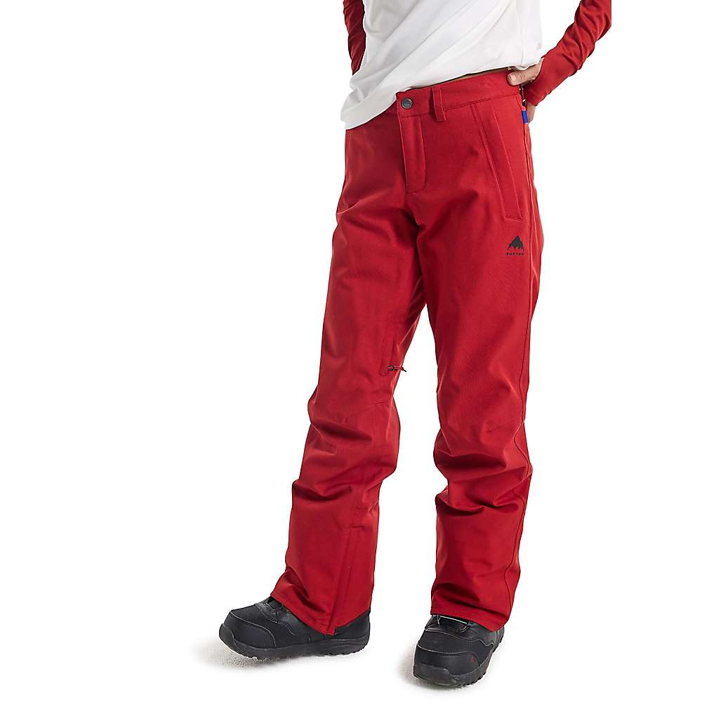 A person in bright red Burton Women's Society Pant, made with DRYRIDE stretch fabric, paired with black snow boots and a white long-sleeve shirt, stands with one hand in their pocket against a plain white background.
