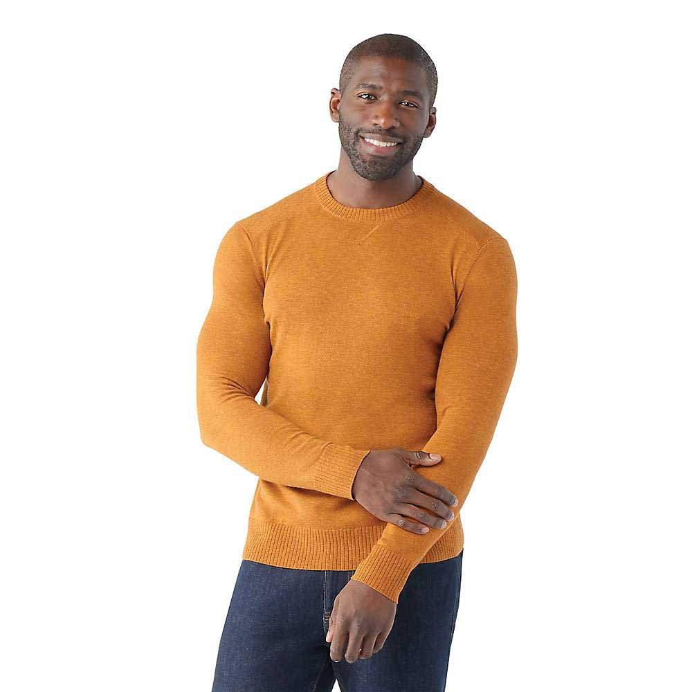 A man in a fitted, long-sleeve, orange Smartwool Mens Sparwood Crew Sweater and dark jeans stands against a white background, smiling and touching his left arm with his right hand.