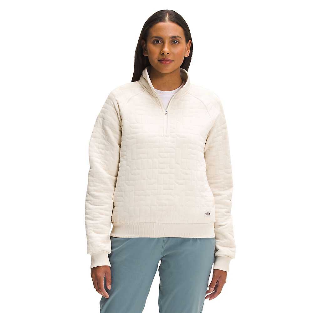A woman stands facing forward, wearing The North Face Women's Longs Peak Quilted 1/4 Zip Jacket in cream and light blue pants against a plain white background.