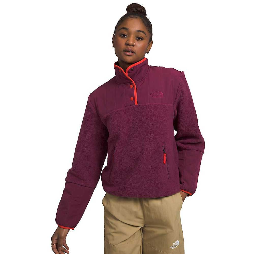 A young woman poses against a white background wearing The North Face Womens Cragmont Fleece 1/4 Snap Top in maroon with red trim and snap buttons, made from recycled polyester, paired with tan pants featuring a white logo.
