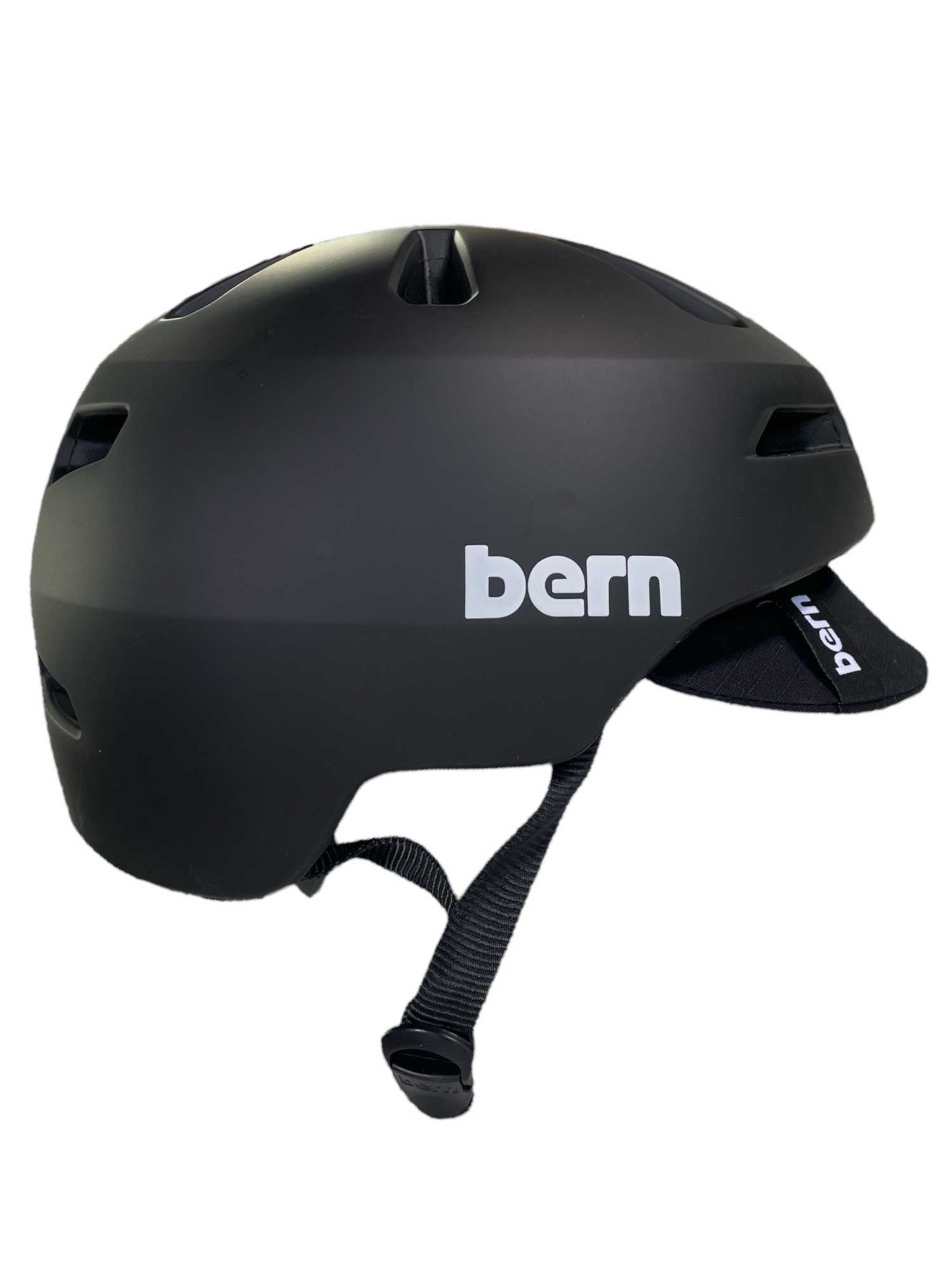 The Bern Brentwood 2.0 Helmet - Bike is a matte black urban helmet with a visor, adjustable chin strap, and visible white Bern logo. Shown from the side, it offers all-season style and safety for every ride.