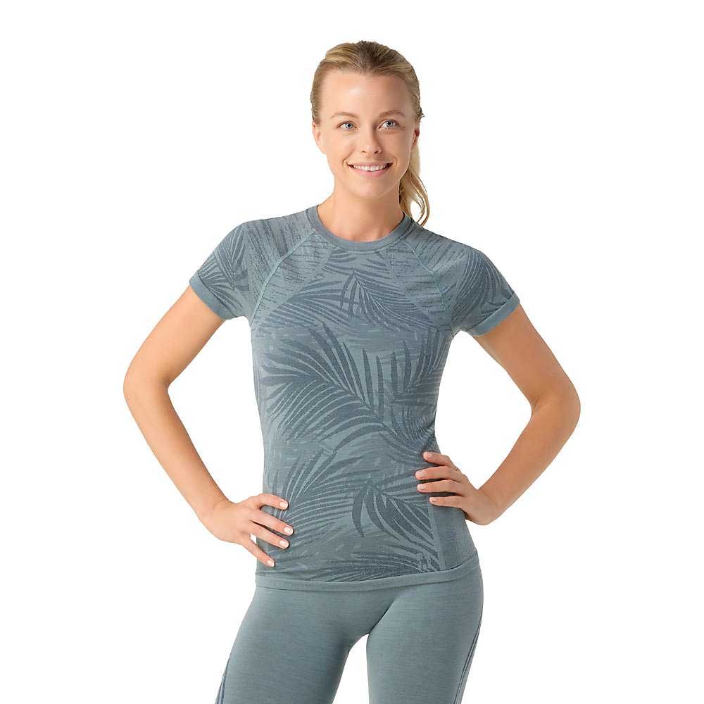 A blonde woman with a ponytail smiles, standing with hands on hips, wearing the blue-green Smartwool Womens Intraknit Active SS Top and matching leggings against a white background.