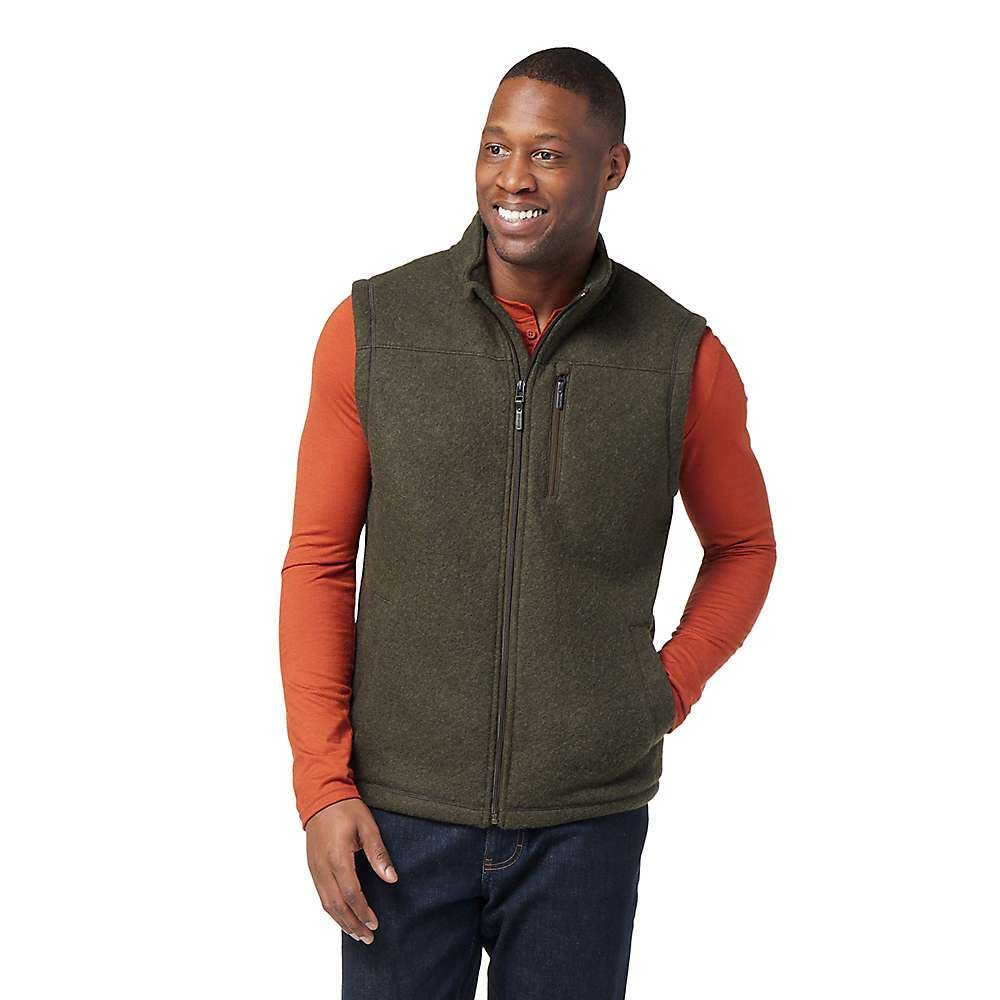 A smiling man stands with one hand in his pocket against a white background, wearing a Smartwool Mens Hudson Trail Fleece Vest made with recycled wool over an orange long-sleeve shirt and dark jeans.