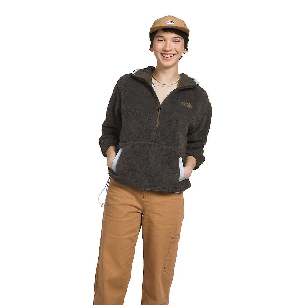 A woman stands smiling with hands in her pockets, wearing The North Face Womens Campshire Fleece Hoodie, tan pants, and a tan cap against a plain white background. The hoodie is made from recycled polyester fleece for warmth and style.