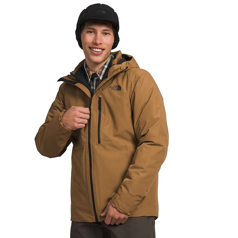 A person wears The North Face Men's North Table Down Triclimate Jacket with a brown hood, a black cap, and a plaid shirt while smiling and adjusting the jacket against a plain white background.