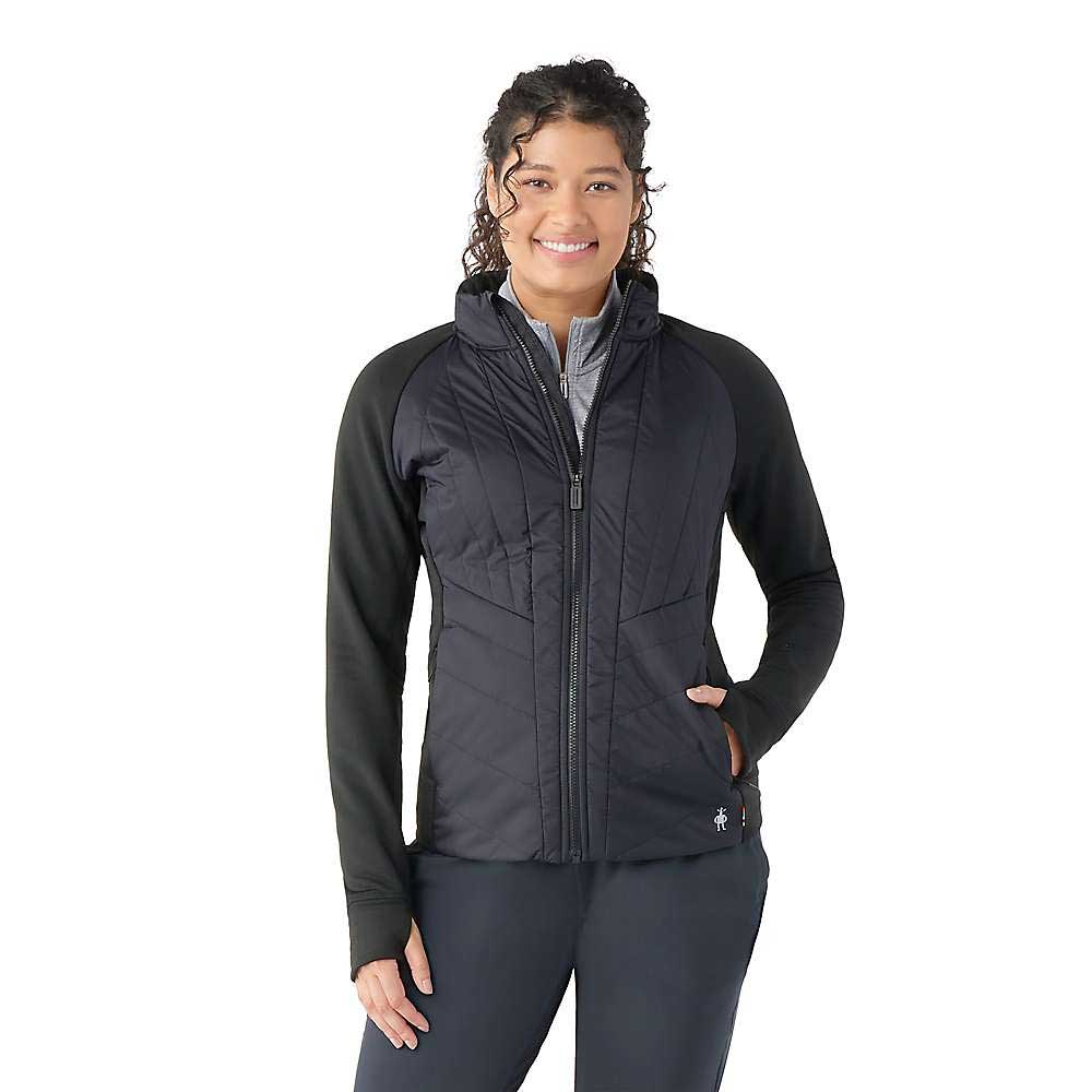 A woman with curly hair smiles while wearing the Smartwool Womens Smartloft Jacket with quilted panels, long sleeves, and Merino wool insulation, standing against a plain white background.