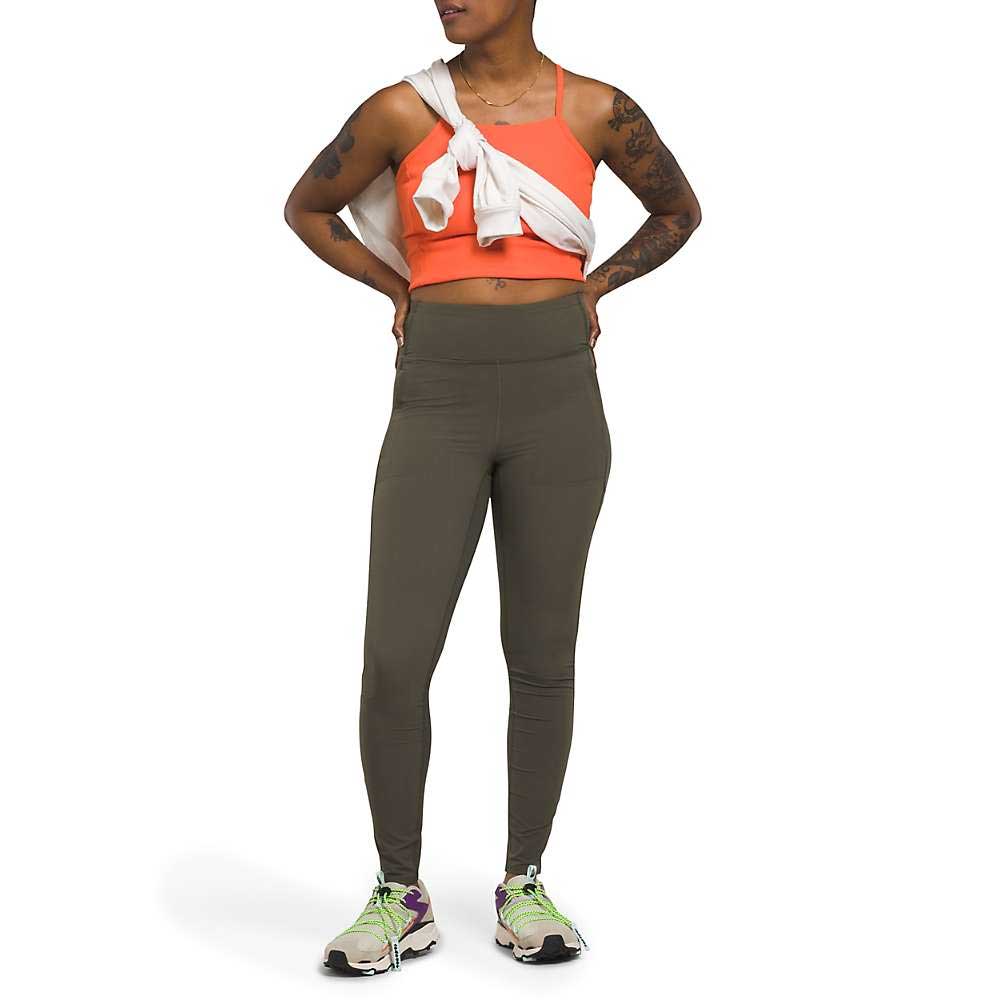 A woman wearing a coral sports bra, The North Face Womens Bridgeway Hybrid Tight in olive, white sneakers with green accents, and a white sweatshirt tied around her shoulders stands with hands on hips, looking to the side.