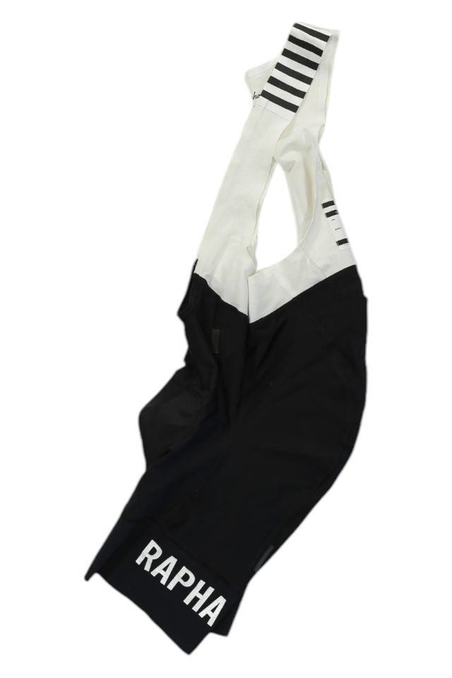A pair of black and white Rapha Mens Pro Team Bib Shorts II - Regular with race-grade chamois, bold RAPHA lettering on the leg, and black stripes on the white shoulder straps.