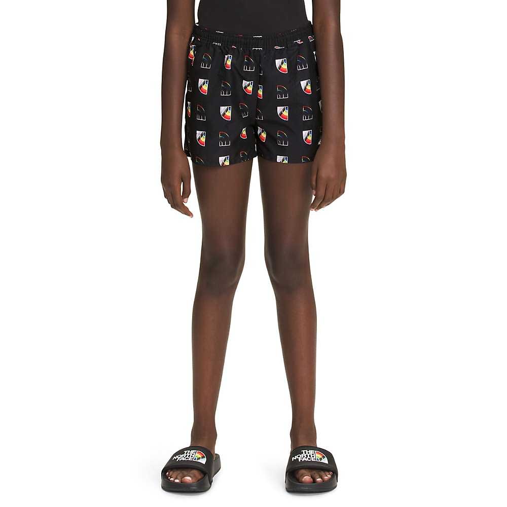 A person wearing a black moisture-wicking T-shirt, The North Face Girls Printed Amphibious Class V Water Short with a colorful logo pattern, and black slip-on sandals stands against a plain white background.