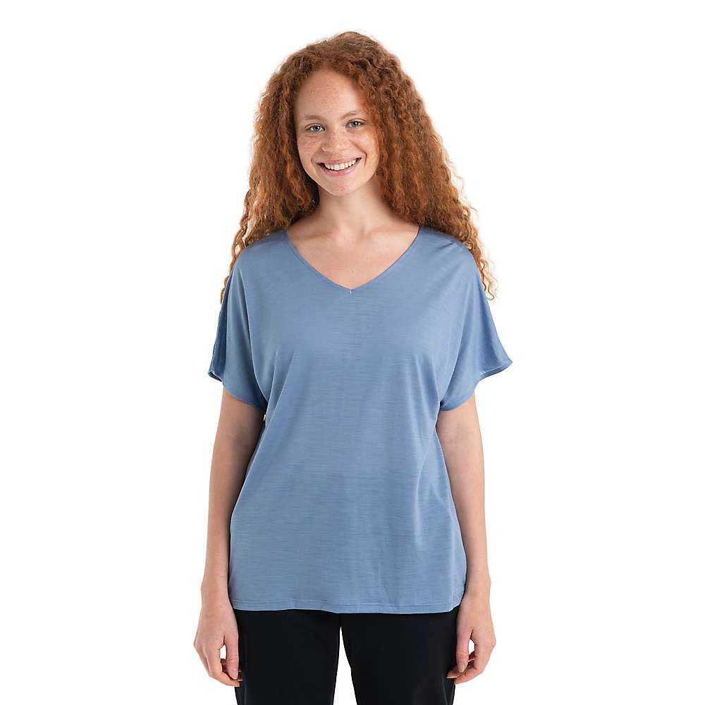 A smiling person with long curly red hair wears the Icebreaker Womens Drayden Reversible SS Top in light blue and black pants, standing against a plain white background.