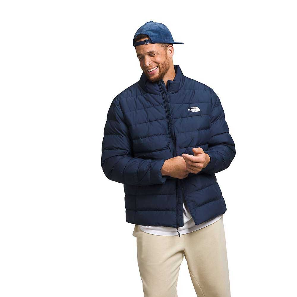 A man wearing The North Face Men's Big Aconcagua 3 Jacket in navy blue, a white shirt, beige pants, and a blue cap smiles while looking down against a plain white background.