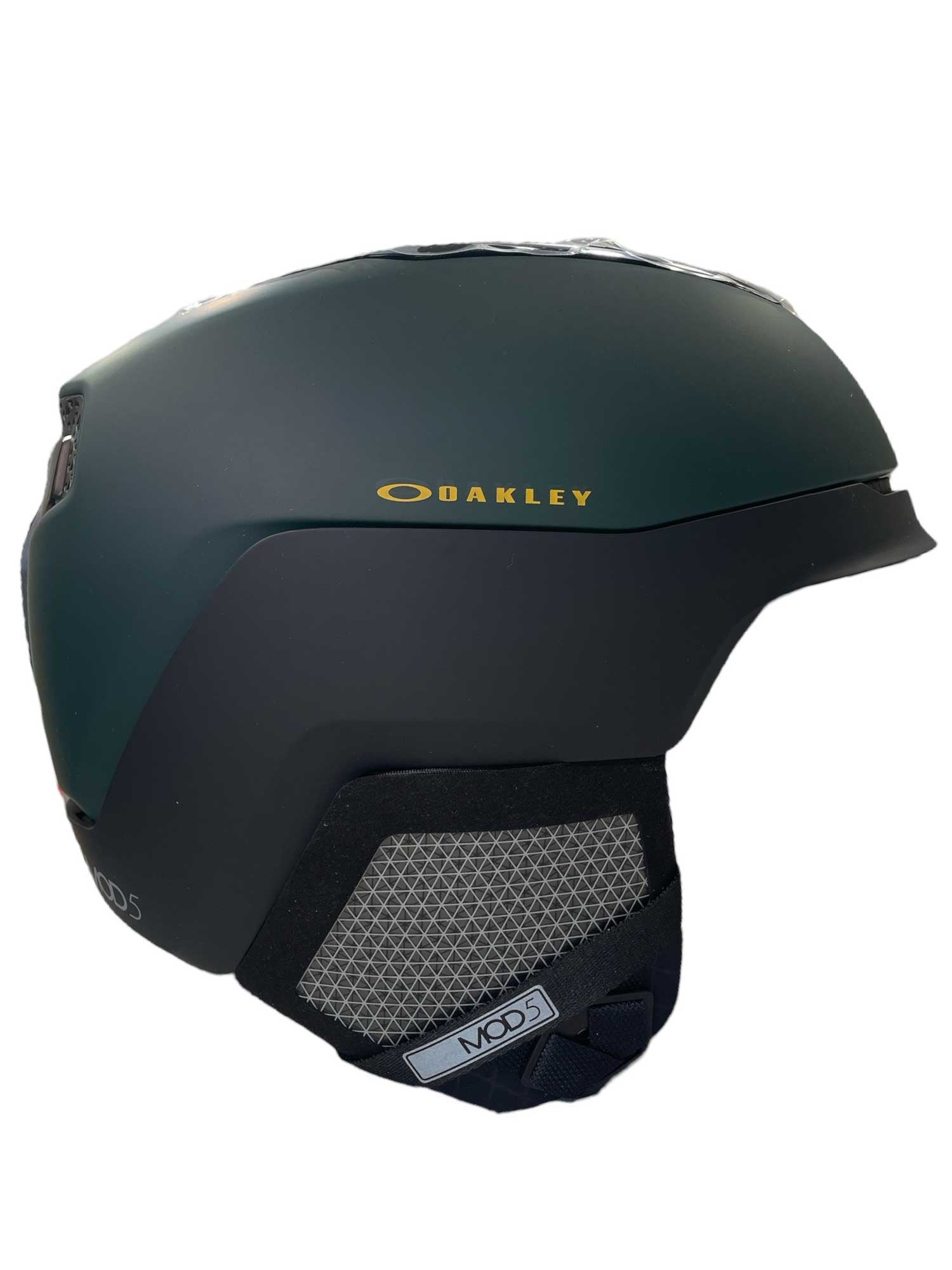 The Oakley Mod 5 Helmet features a matte dark green finish with black and gray ear coverings, mesh vent with adjustable ventilation, and seamless helmet-goggle integration. A yellow Oakley logo stands out on the side.