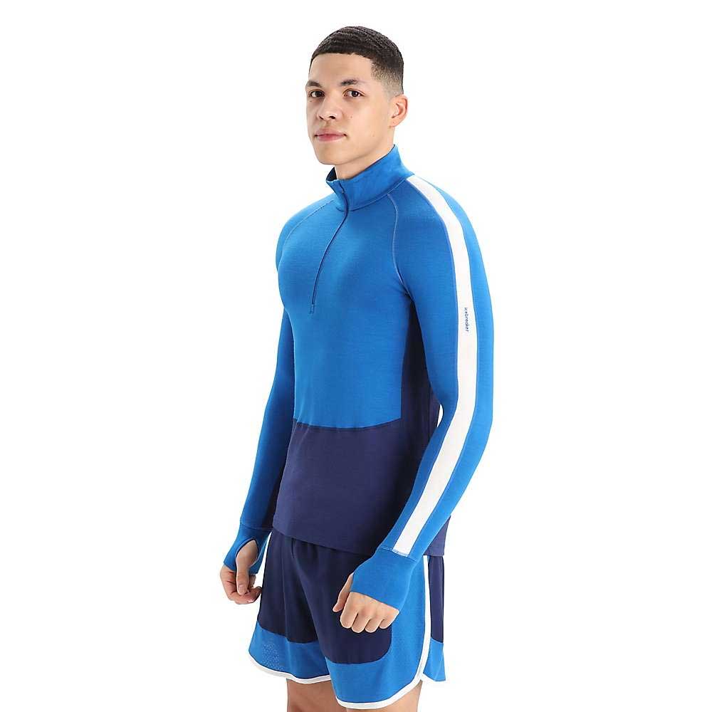 A young man stands against a plain white background, wearing the Icebreaker Mens 260 Zoneknit LS Half Zip Top in blue with a zip collar and white sleeve stripe, paired with blue running shorts.