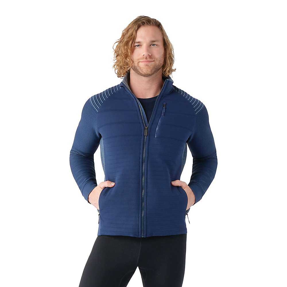 A person with wavy shoulder-length hair wears the Smartwool Men's Intraknit Merino Insulated Jacket in blue, featuring horizontal stitching and pockets, standing with hands in pockets against a white background.