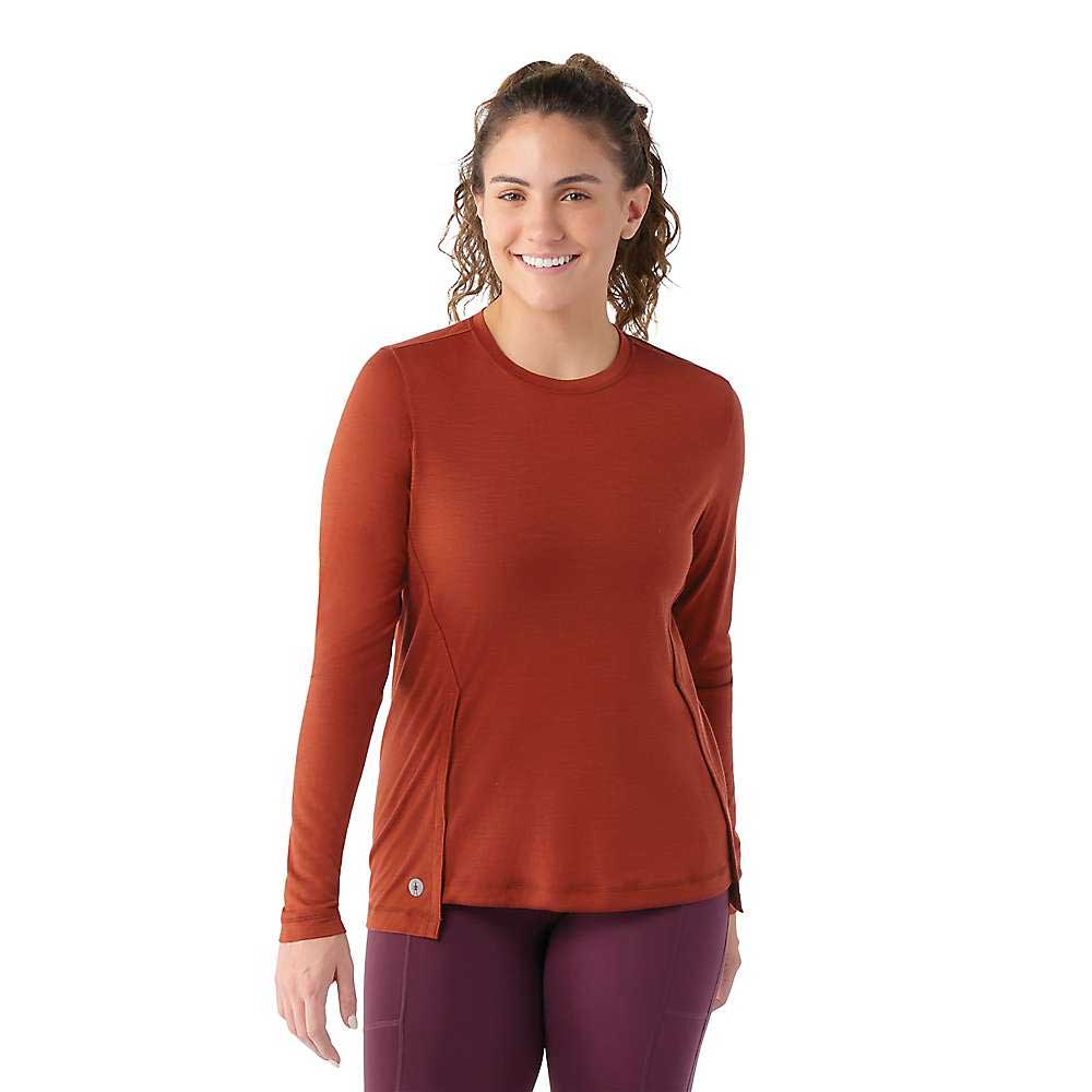 A woman with curly hair, wearing the Smartwool Womens Active LS Crew Top in rust and maroon leggings, smiles against a white background.