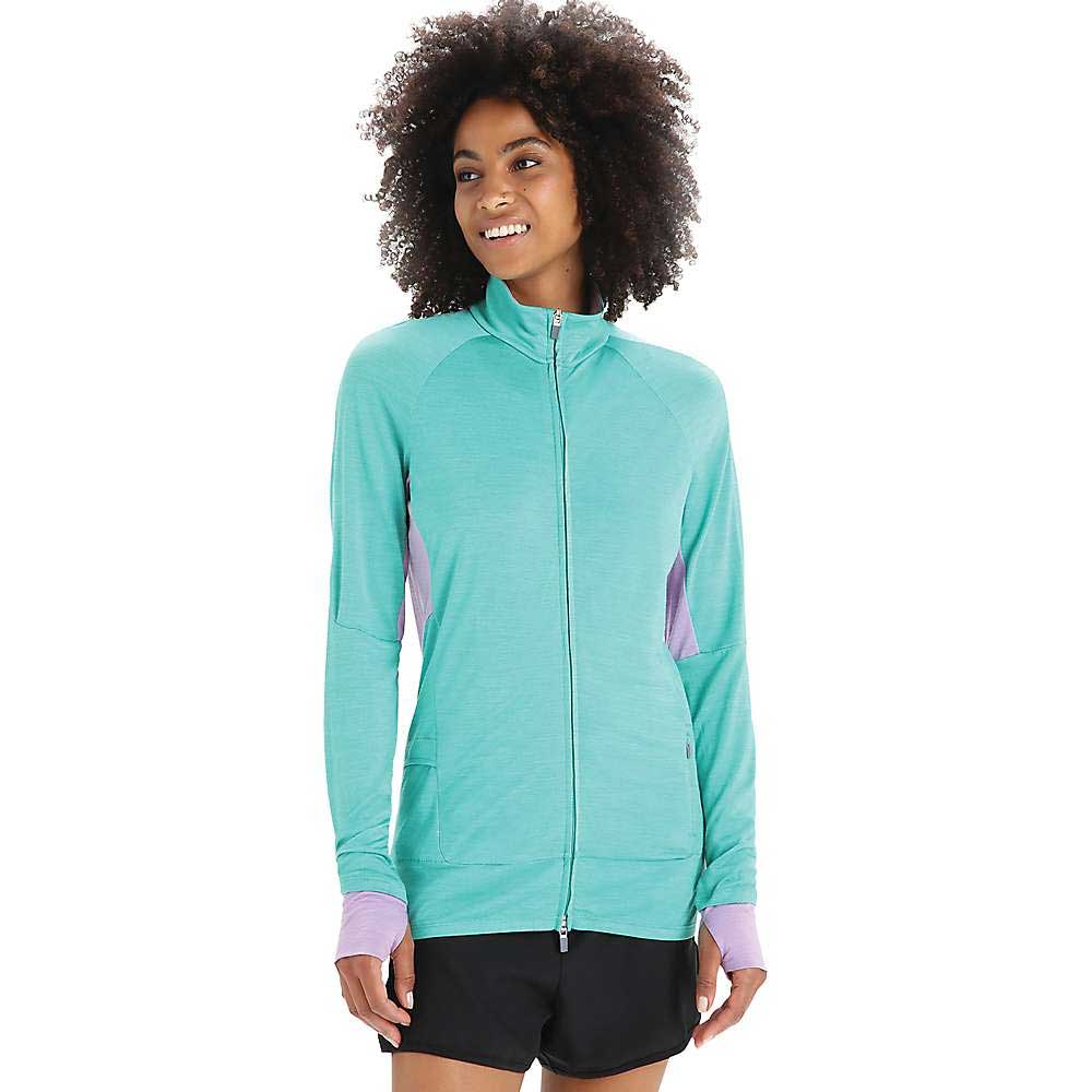 A woman with curly hair wears the Icebreaker Womens ZoneKnit LS Zip Jacket in turquoise with lavender cuffs and sides, paired with black shorts. She stands smiling, looking slightly to the side against a plain white background.