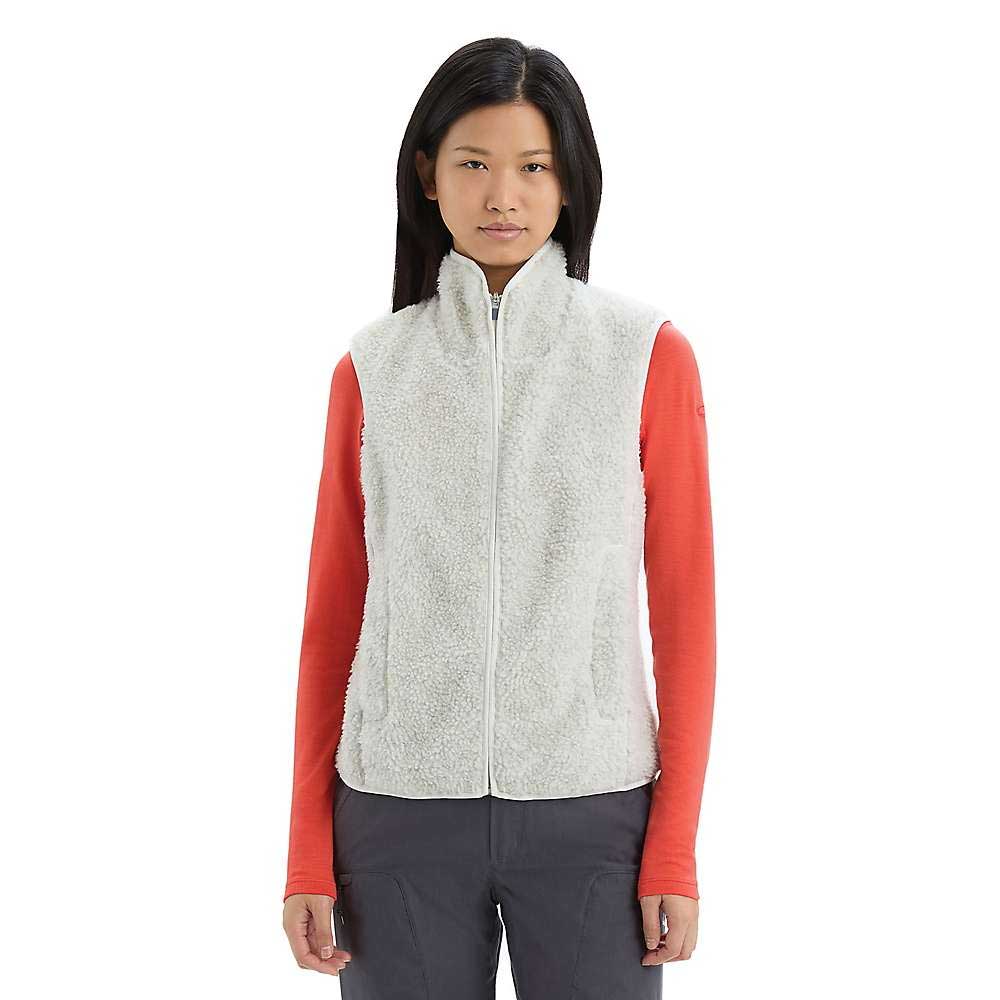 A woman faces forward, wearing the Icebreaker Women's RealFleece High Pile Vest in white sherpa over a long-sleeve red shirt and gray pants, set against a plain white background.