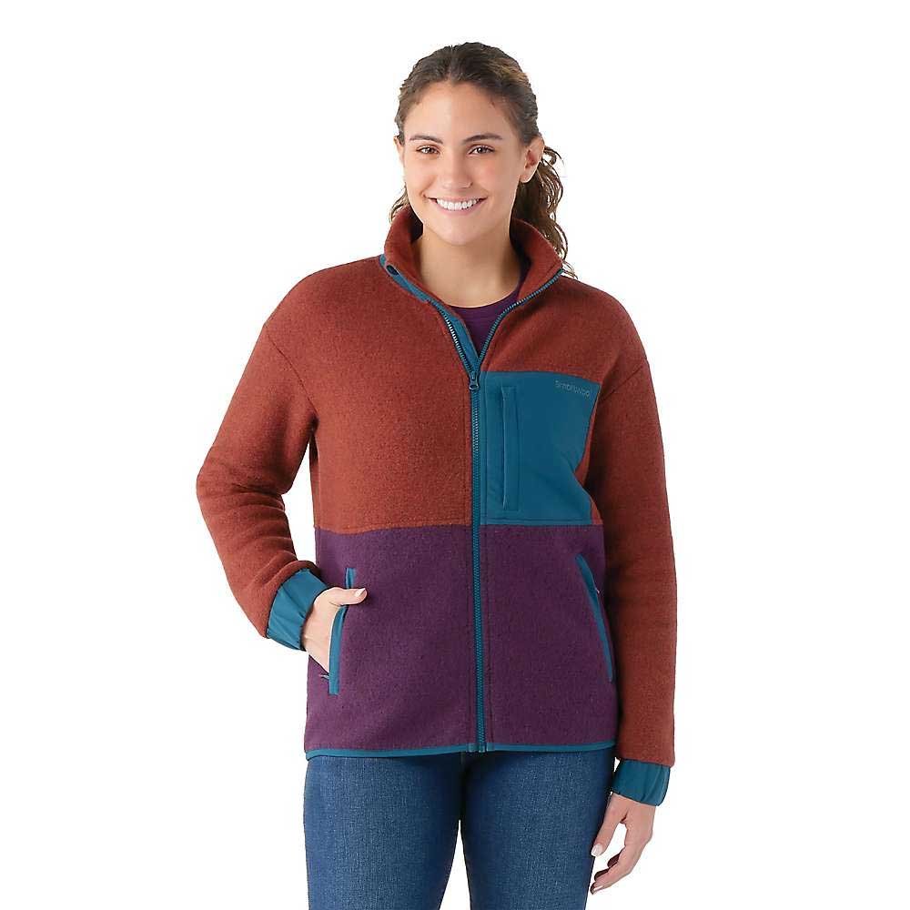 A woman smiles in the Smartwool Womens Hudson Trail Fleece Jacket in rust, purple, and teal with zippered pockets, made from recycled wool fleece, standing against a plain white background.
