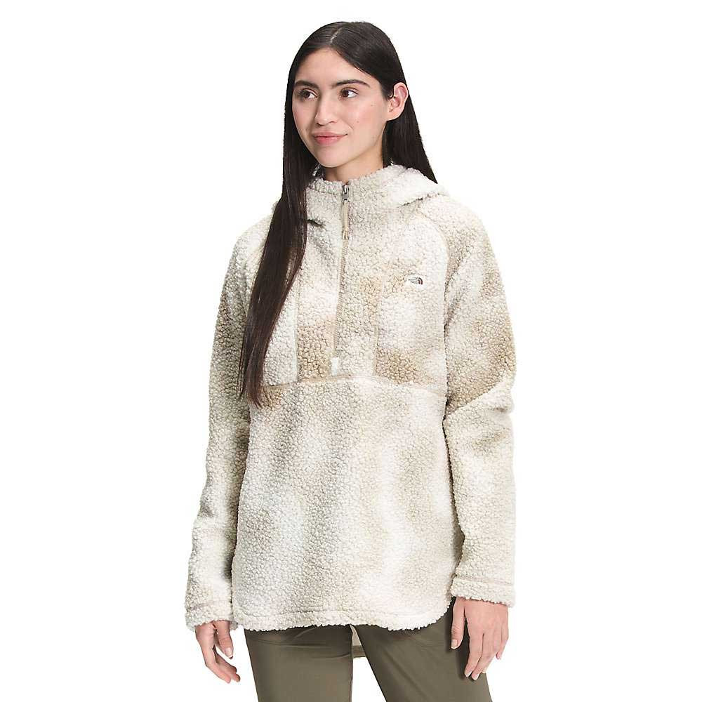A woman with long dark hair models The North Face Womens Printed Ridge Fleece Tunic, a cream and tan Sherpa pullover hoodie with a half-zip and small chest logo, standing against a plain white background.